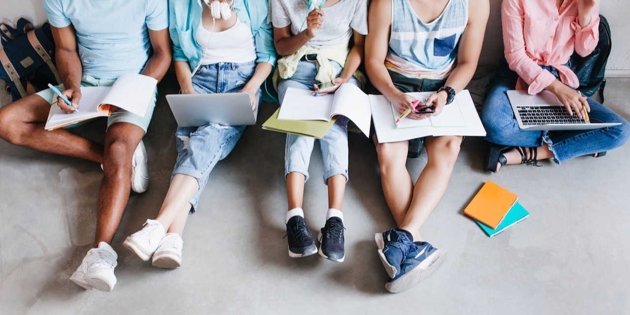 Overhead portrait of young people with laptops and smartphones, sitting together on the floor. Students writing lectures holding textbooks on their knees..